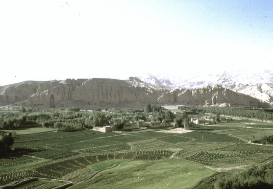 A panoramic view of green fields, trees and houses, and large stone cliffs with mountains beyond.