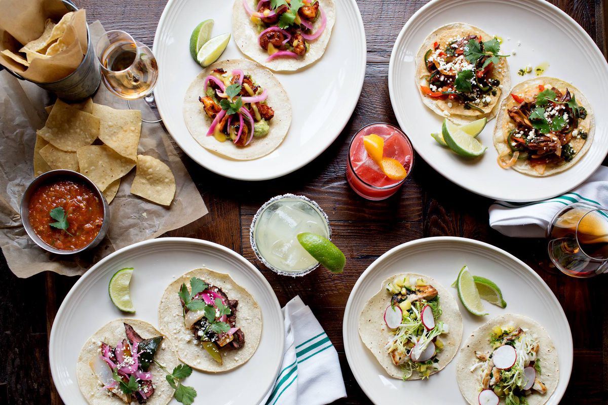 Overhead view of four plates of tacos, plus chips and salsa and cocktails.