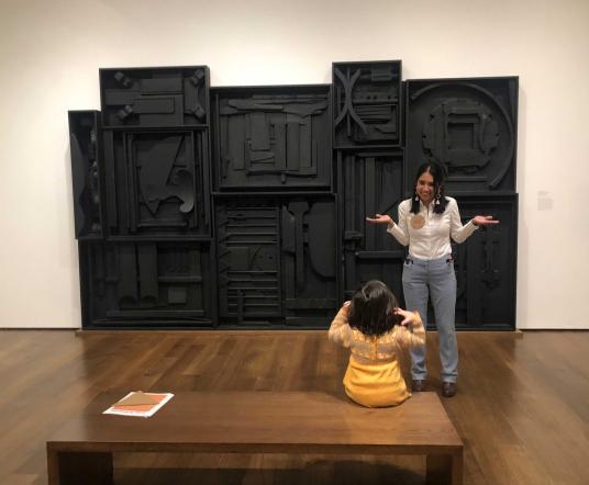 A woman shrugs her shoulders in front of a black sculpture; a young girl copies her motion.