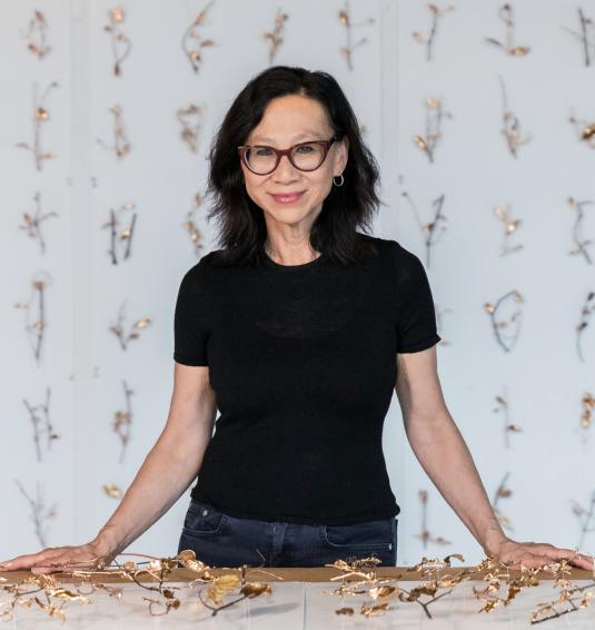 A woman with dark hair and glasses poses in front of a table with gold leaf vines.