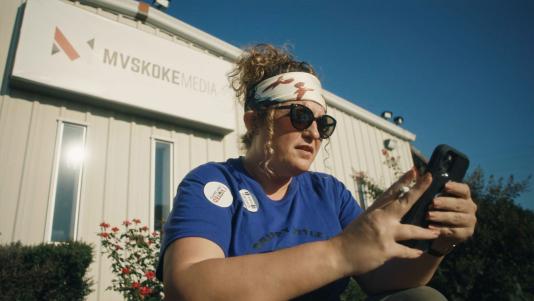 A woman looking at her phone sits in front of a building that has a sign reading “Mvskoke Media.”