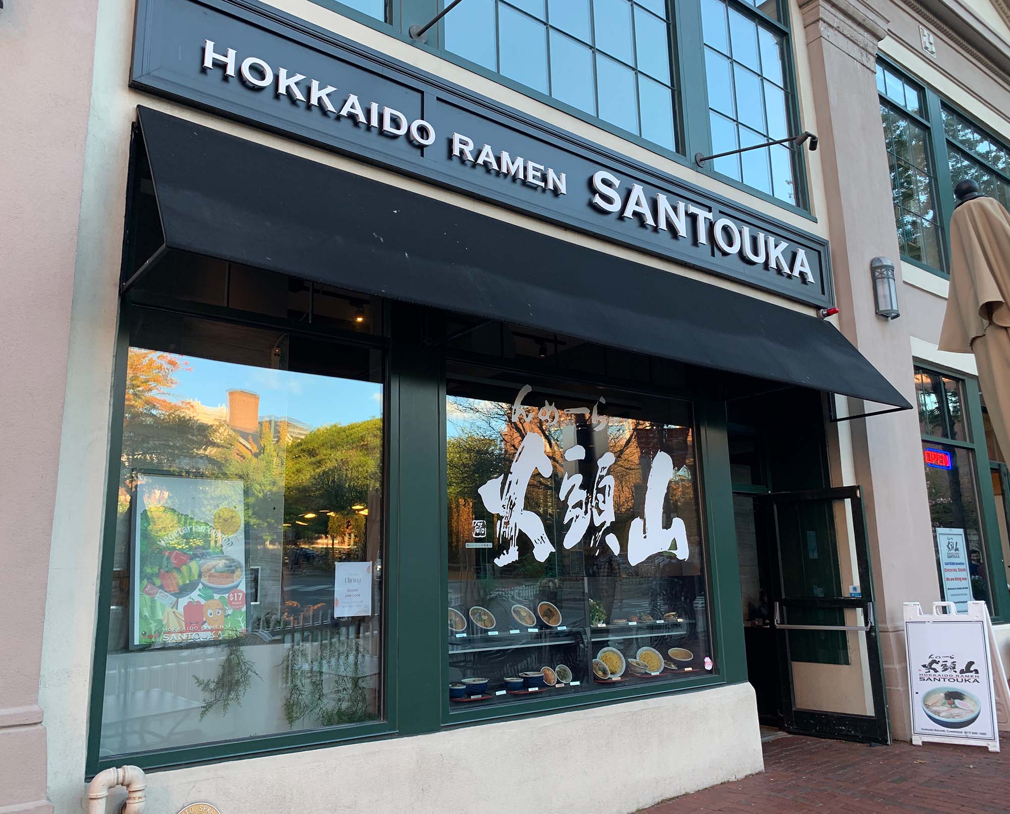 Photo taken outside Hokkaido Ramen Santouka. The facade has dark green trim and has Japanese characters in the window painted in white, as well as a menu and ramen models on display.