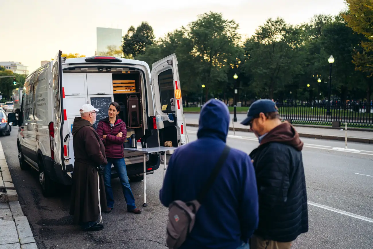 People gather near a Capuchin Mobile Ministries van parked on a city street with open doors.