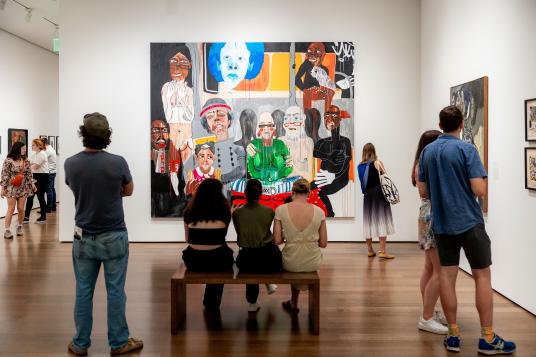 Museum visitors observe various artworks, including a large, colorful painting featuring abstract and figurative elements. Some people stand while others sit on a bench.