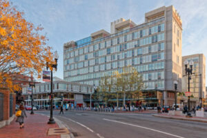 Modern glass-fronted building on a city street with pedestrians, bicycles, autumn trees, and streetlamps