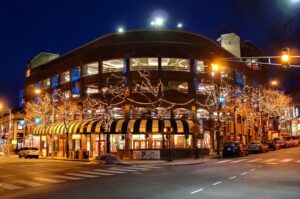 Illuminated multi-story commercial building with storefronts and tree lights at a city intersection at night